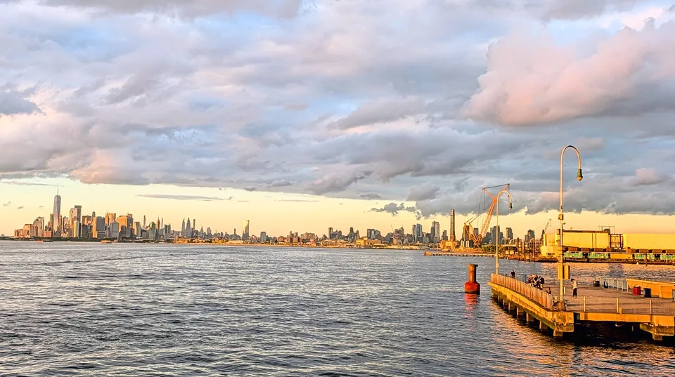 The Manhattan skyline across the river, with a sunlit dock and pink clouds.