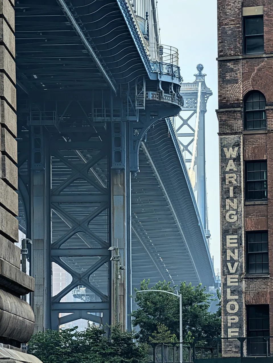 The underside of the Manhattan Bridge framed by brick buildings and trees.