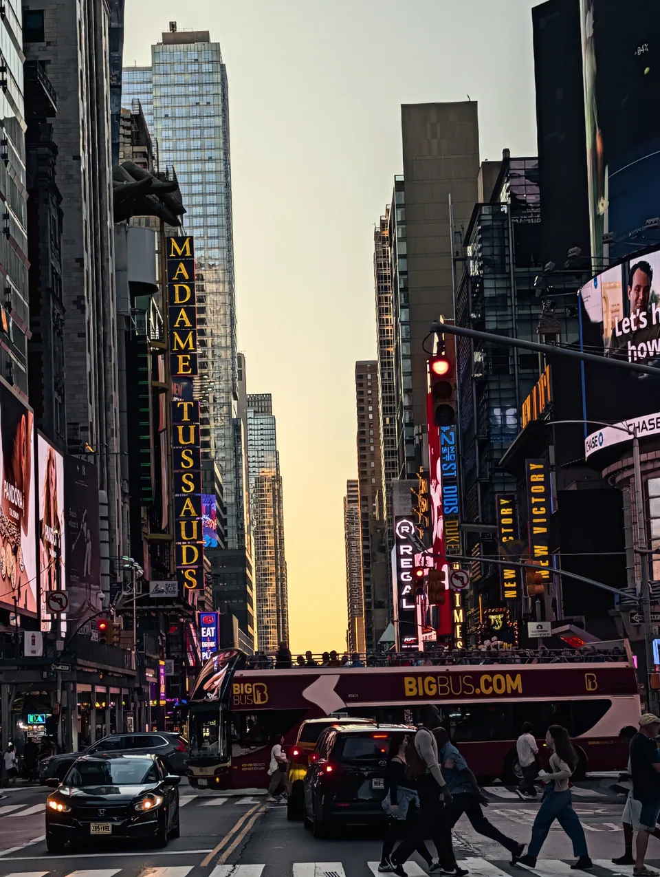People crossing a busy New York street between towers and illuminated signs at sunset.