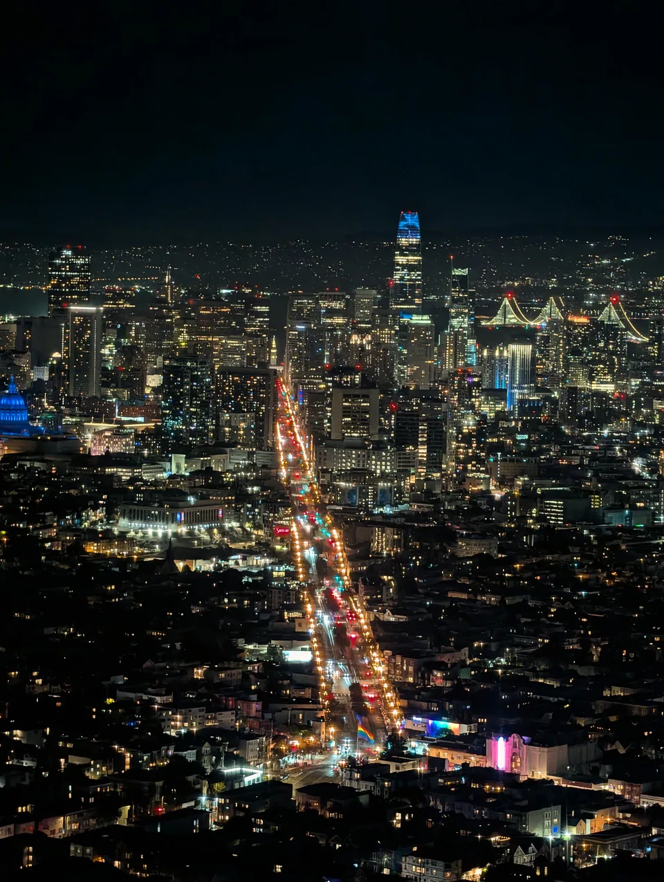 San Francisco and the illuminated Bay Bridge at night, with Market Street leading towards a blue-lit Salesforce Tower.