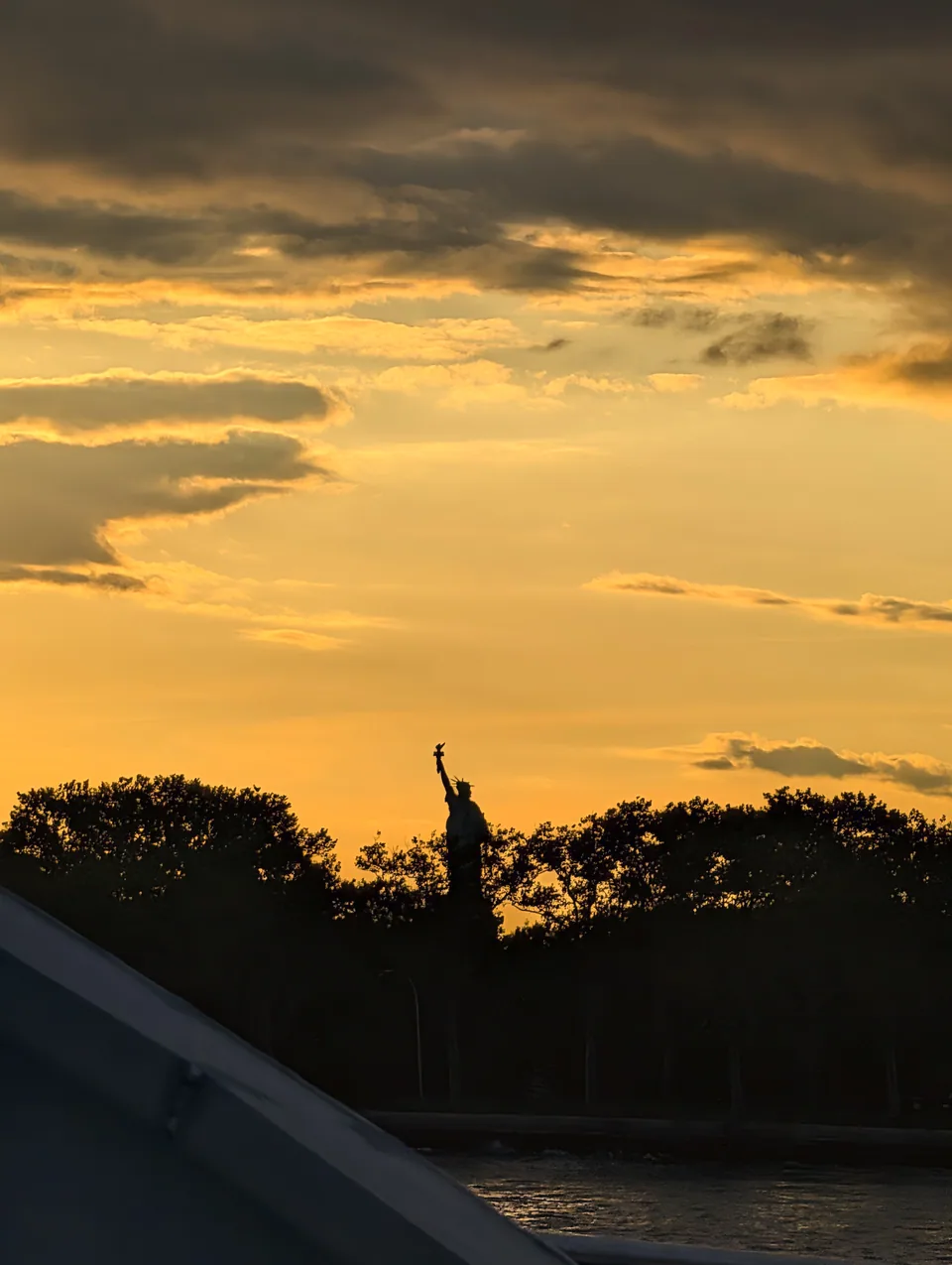 The Statue of Liberty silhouetted behind trees under a golden evening sky.