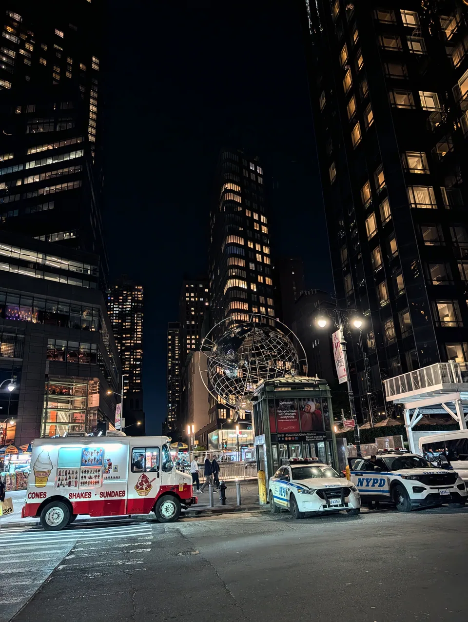 An illuminated globe, food trucks and buildings at Columbus Circle at night.
