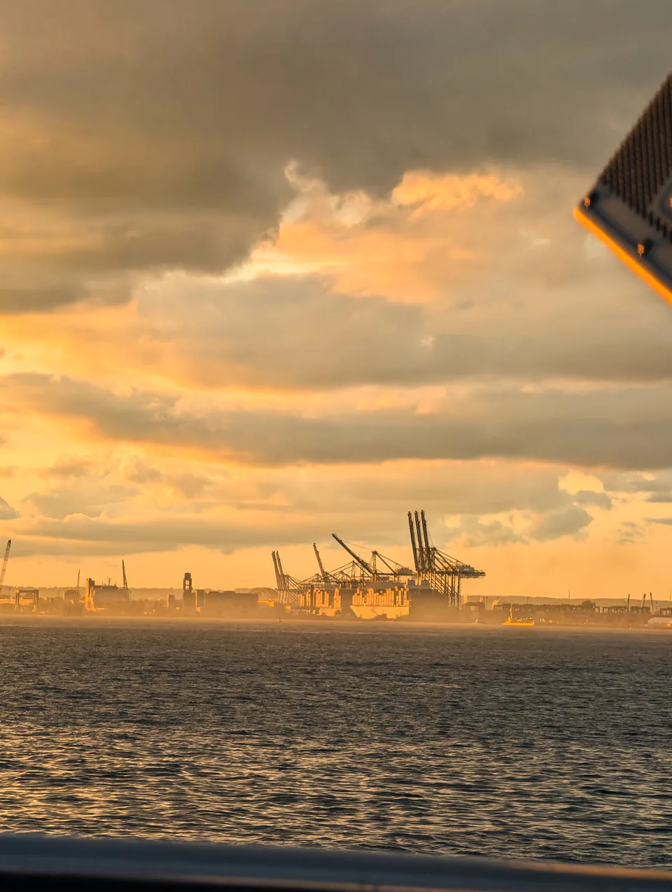 Port cranes along a hazy shore beneath sunset clouds, seen from a ferry.