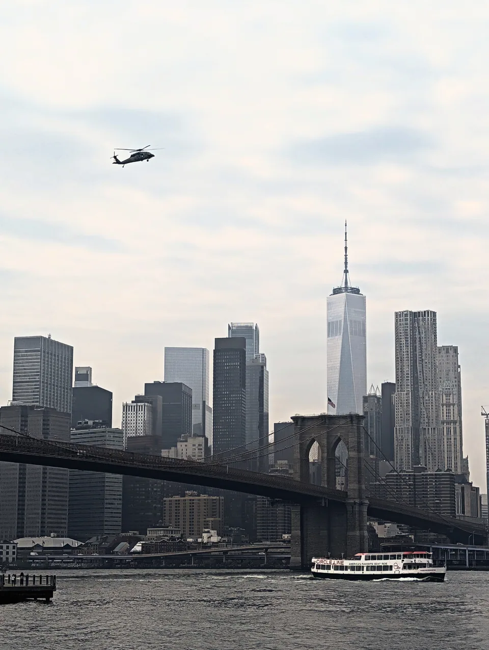 The Manhattan skyline and Brooklyn Bridge beneath a cloudy sky, with a helicopter overhead.